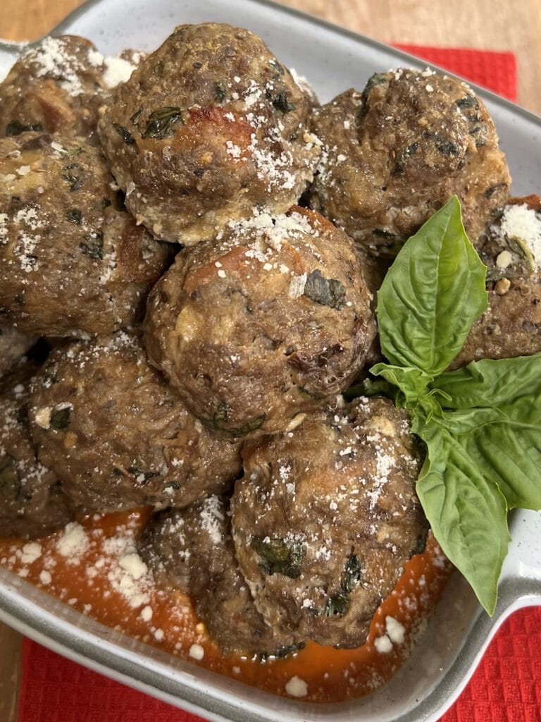 Eggplant and beef meatballs sprinkled with grated cheese and basil garnish over tomato sauce in white serving dish.