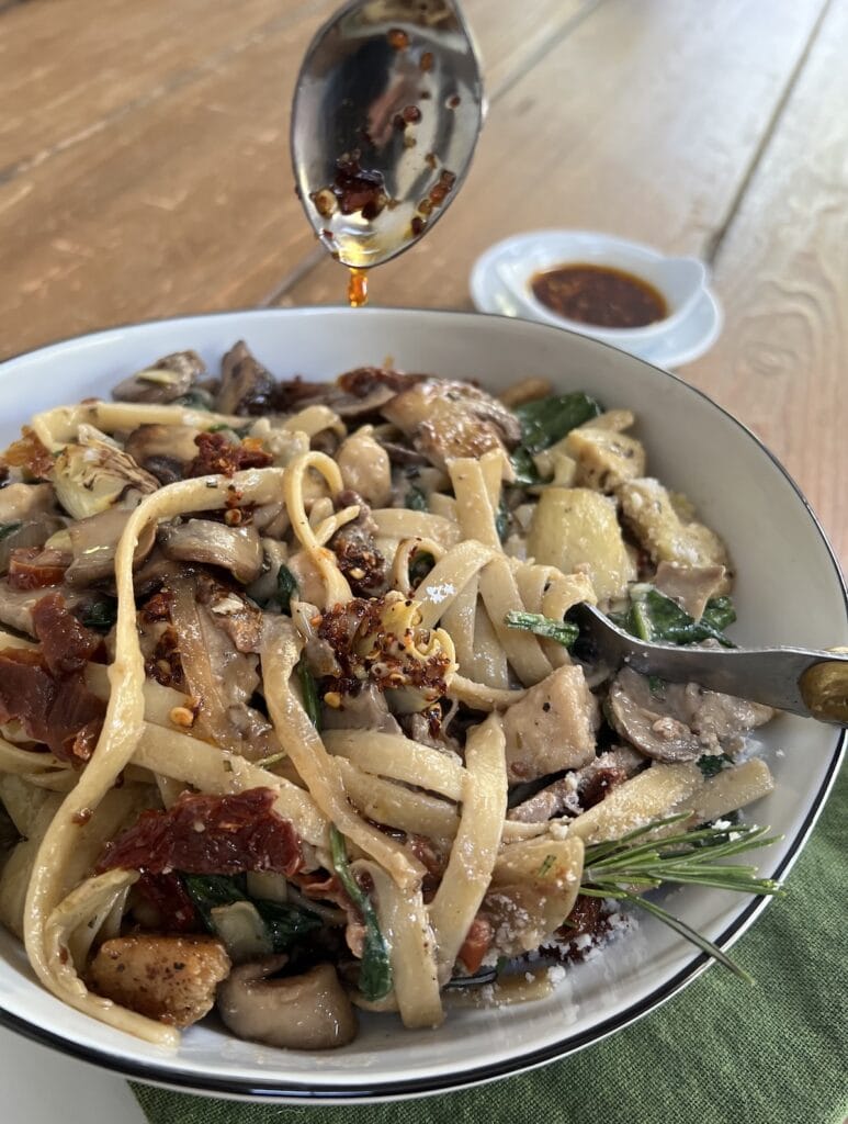 Drizzling homemade olio santo into bowl of Chicken Marsala Noodles with spoon.