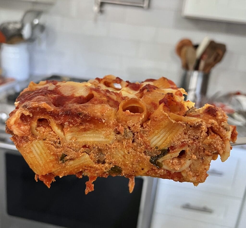 Side view of a large slice of pasta al forno on a spatula with kitchen in the background.