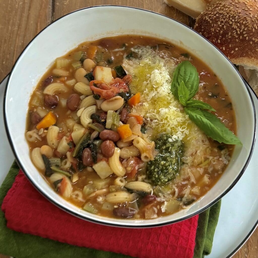 Italian minestrone soup in white soup bowl with bread in background.