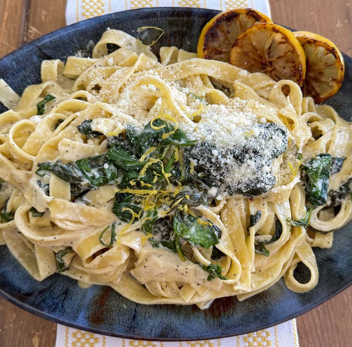 Creamy lemon pasta with spinach in blue serving platter with sliced lemons and lemon zest garnish and sprinkled with grated pecorino cheese.