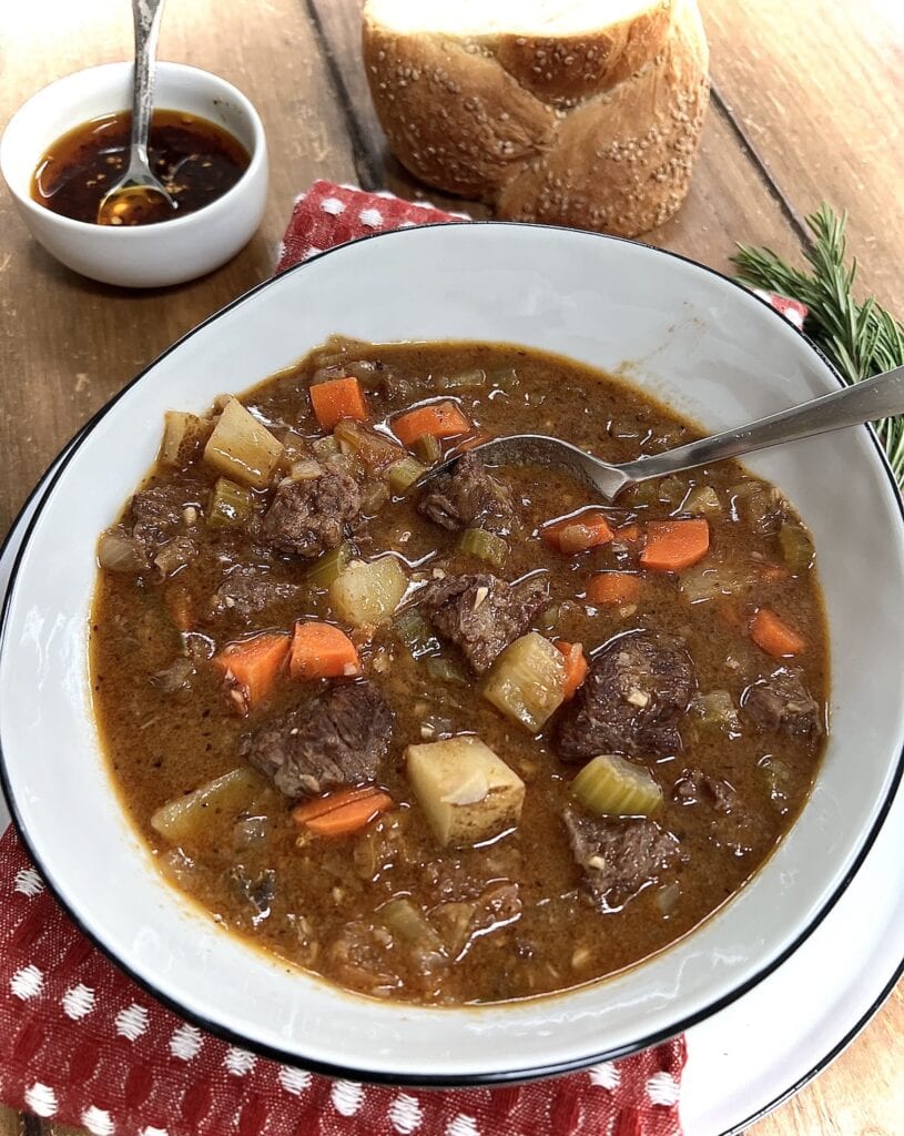 Italian Beef Soup in white soup bowl with spoon and spicy oil and bread in background.
