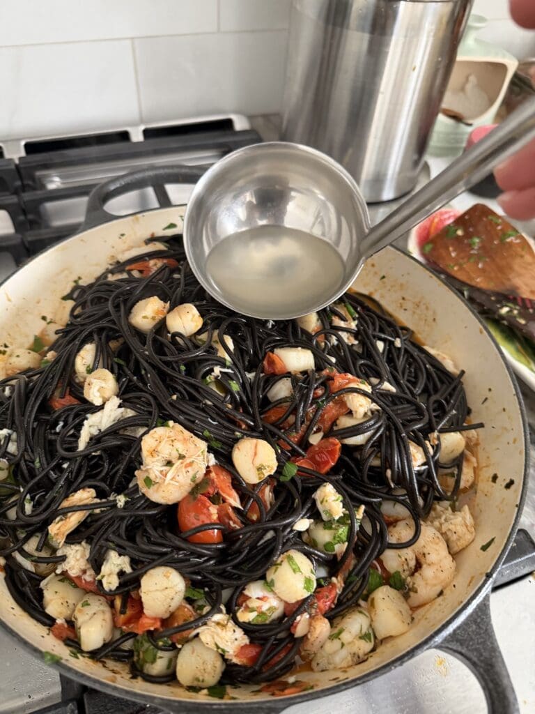 Adding some pasta water to Squid Ink Pasta With Seafood in pan for additional mositure.