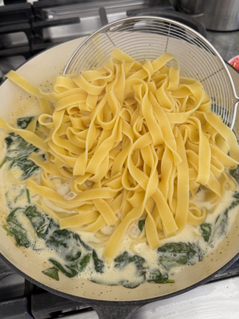 Adding cooked tagliatelle pasta to creamy spinach sauce to let it finish cooking in the sauce.