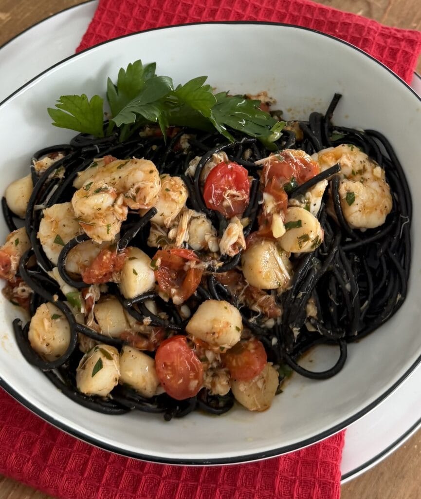 Squid Ink Pasta With Seafood in white pasta bowl with parsley garnish over decorative red towel.