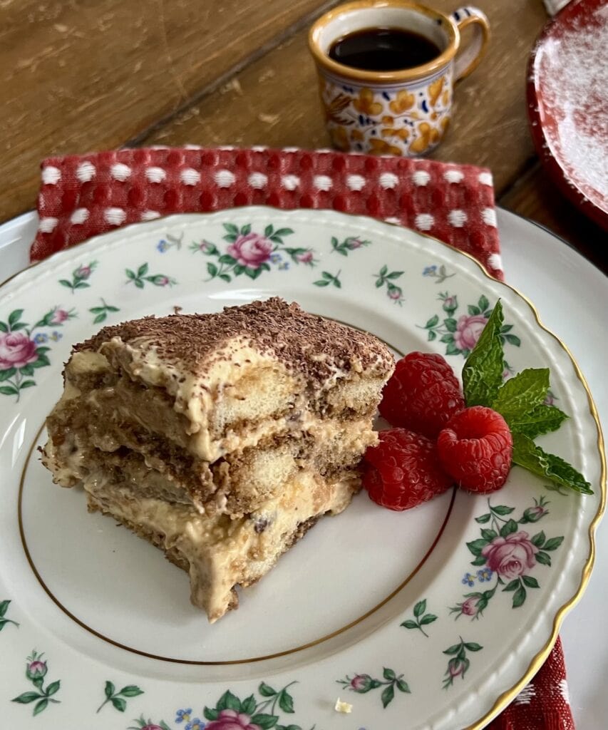 A slice of Classic Italian Tiramisu Recipe with raspberry garnish on decorative china plate with cup of espresso in the background.