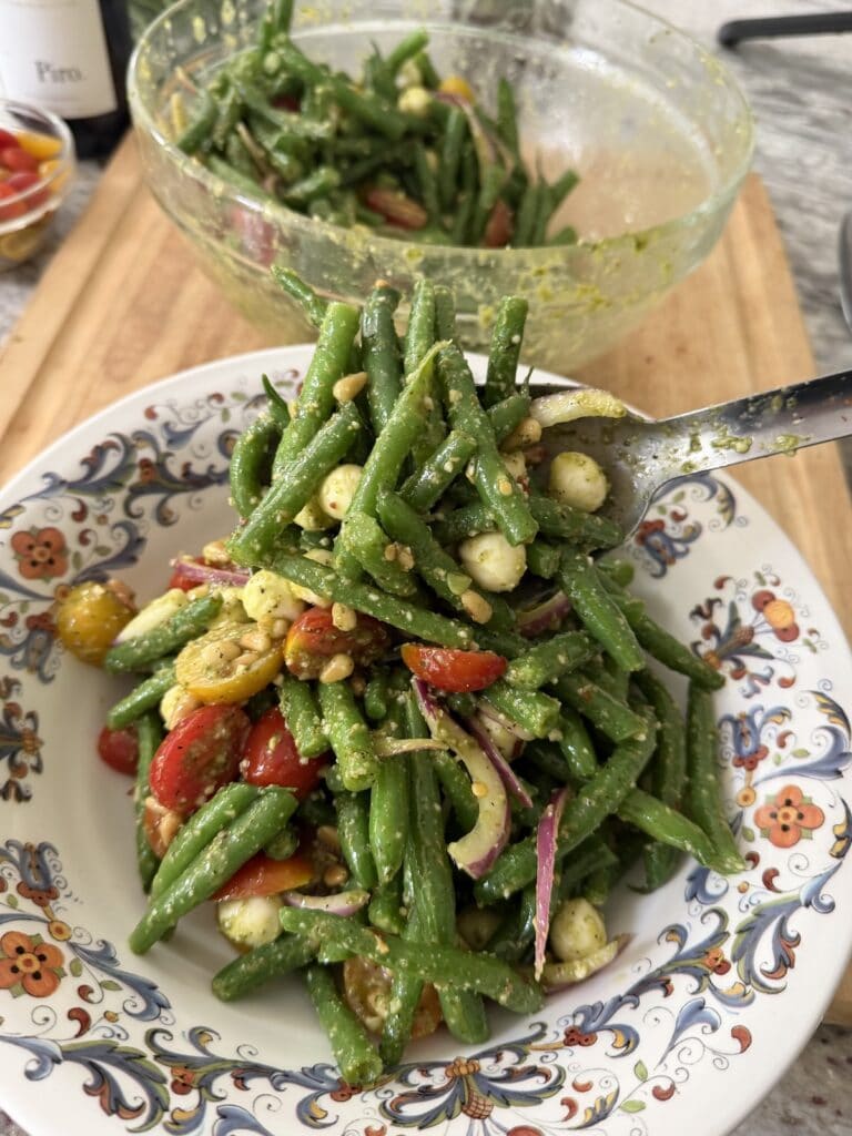 Spooning Italian Green Bean Salad into serving bowl from mixing bowl in background.