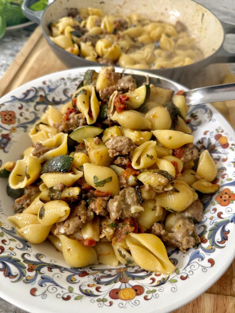 Spooning Zucchini and Sausage Pasta into decorative bowl from pan in background.