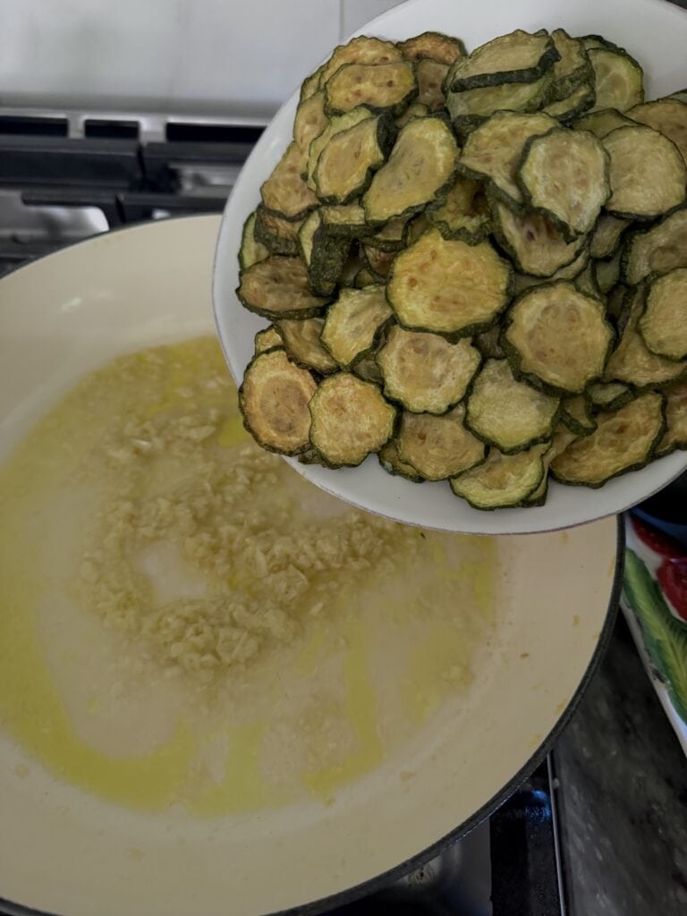 Adding rested fried zucchini slices to garlic in pan.