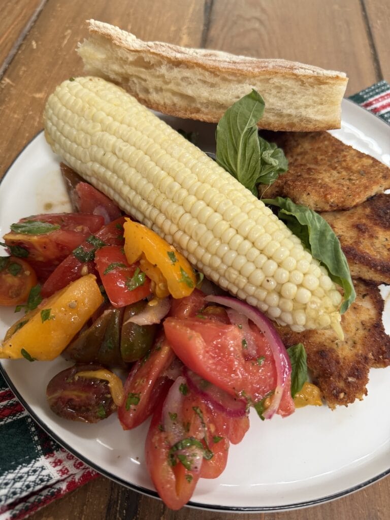 Italian marinated tomatoes on dinner plate with corn, bread and chicken cutlets.