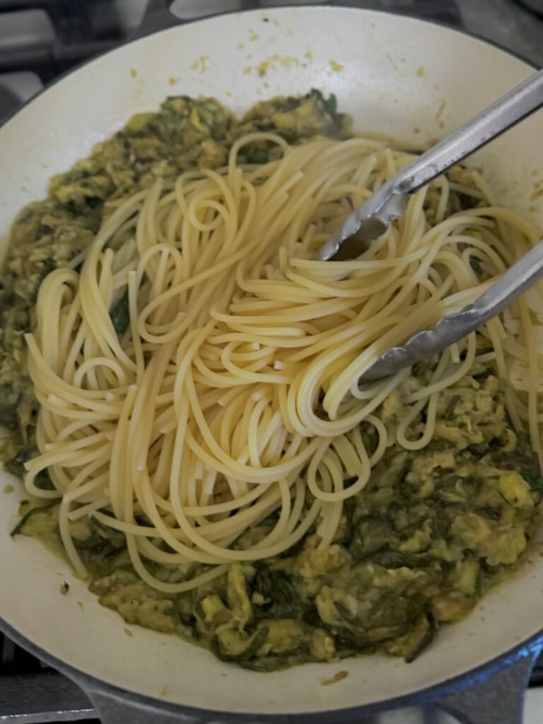 Undercooked spaghetti pasta just added to zucchini sauce in pan with tongs.