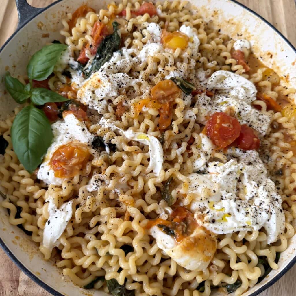 Burst tomato pasta with burrata with basil garnish in pan.
