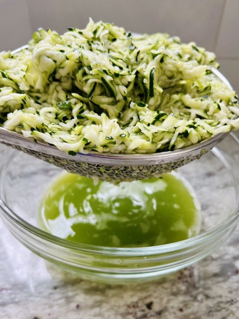 Shredded zucchini in a colander over a bowl with residual liquid that has been squeezed out of it.