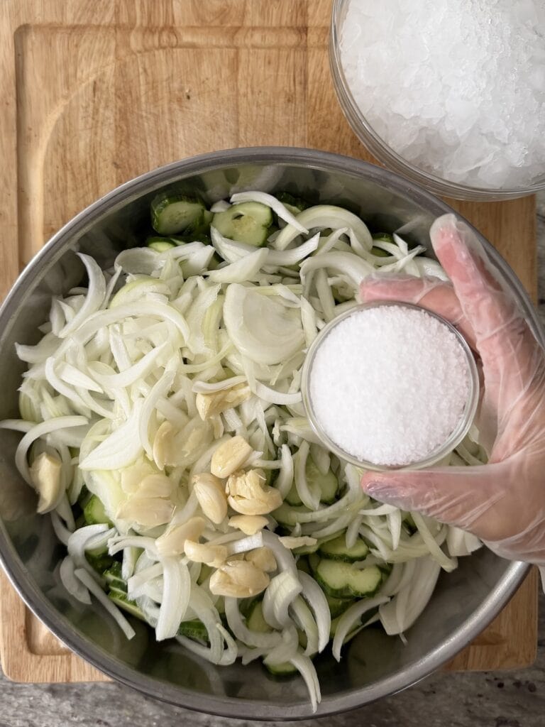 Bowl of salt about to be added to raw sliced cucumber, onion, garlic and salt mixture in bowl.