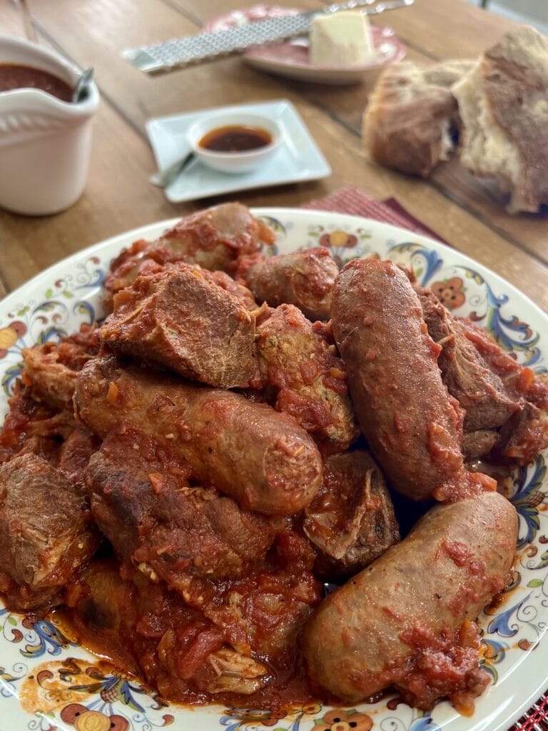 Bowlful of braised meats in sauce with bread, cheese, sauce and spicy oil in background.