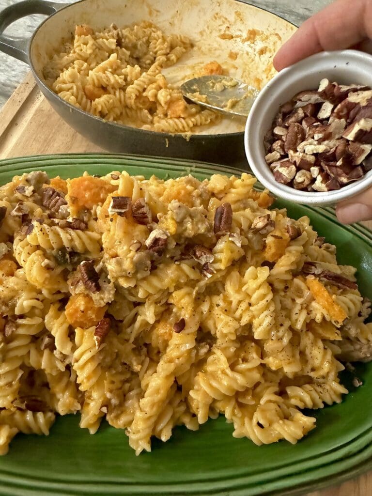 Sprinkling toasted chopped pecans over finished creamy fall pasta in serving platter.
