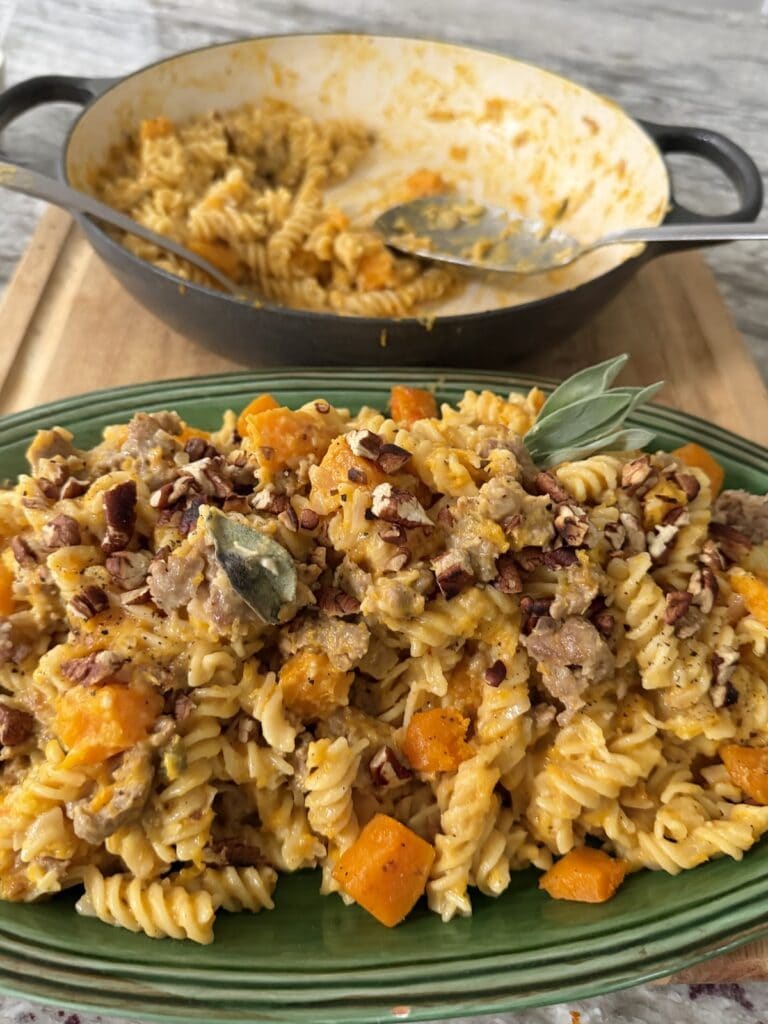 Creamy Butternut Squash and Sausage Pasta on green serving platter with sage garnish, with pan of it in the background with spoons
