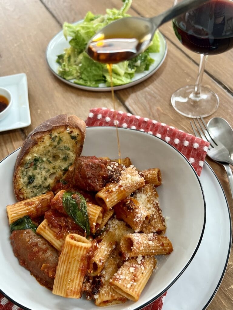 Drizzling Italian chili oil (olio santo) over bowl of Sunday sauce for a little heat, with salad and glass of red wine in background.