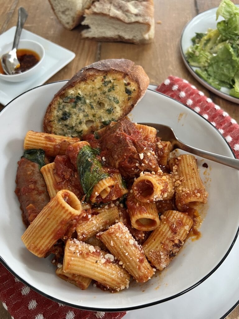 Pasta bowl filled with Italian sunday Gravy and garlic bread with fork and salad and spicy oil in background.