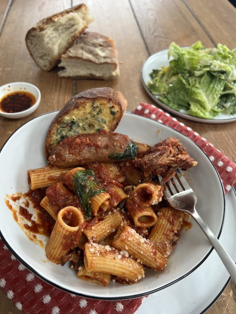Best Italian Gravy Recipe in pasta bowl with for, and garlic bread, with spicy oil, bread and caesar salad in background.