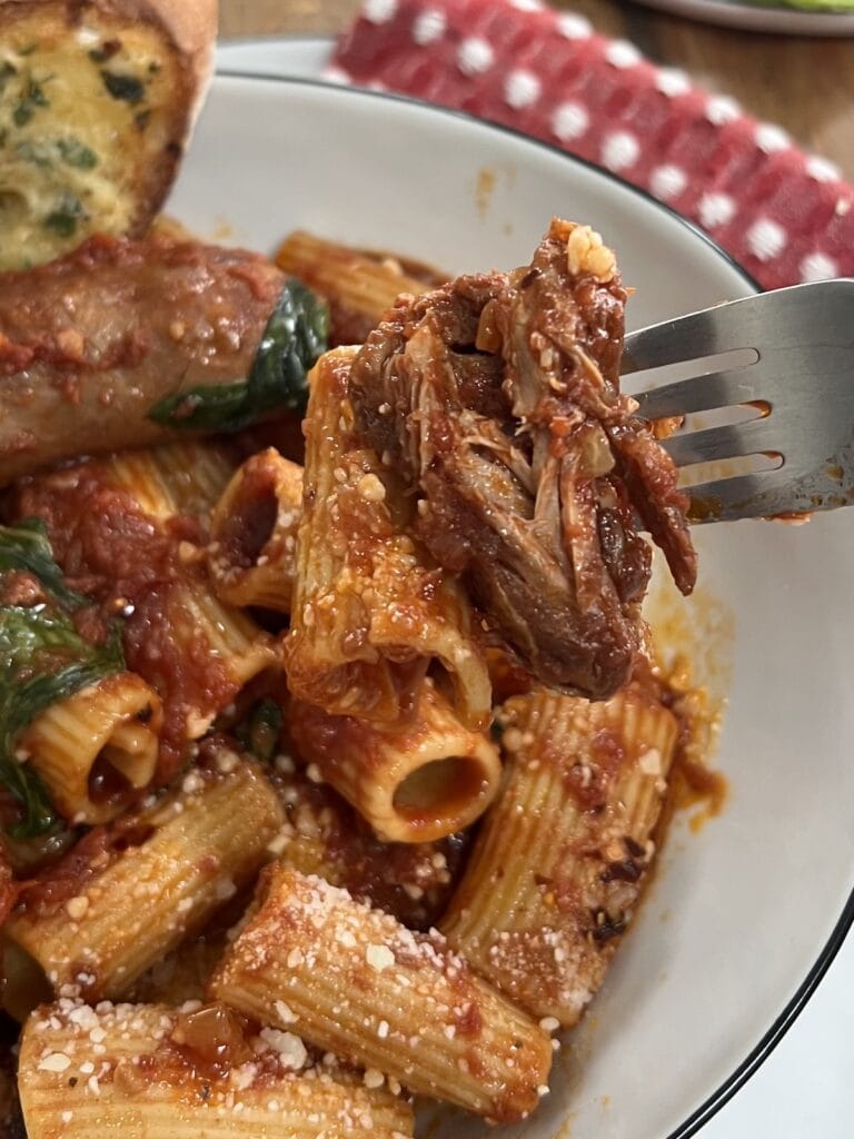 Close up of a pice of braised meat on fork over a bowl of macaroni and gravy.