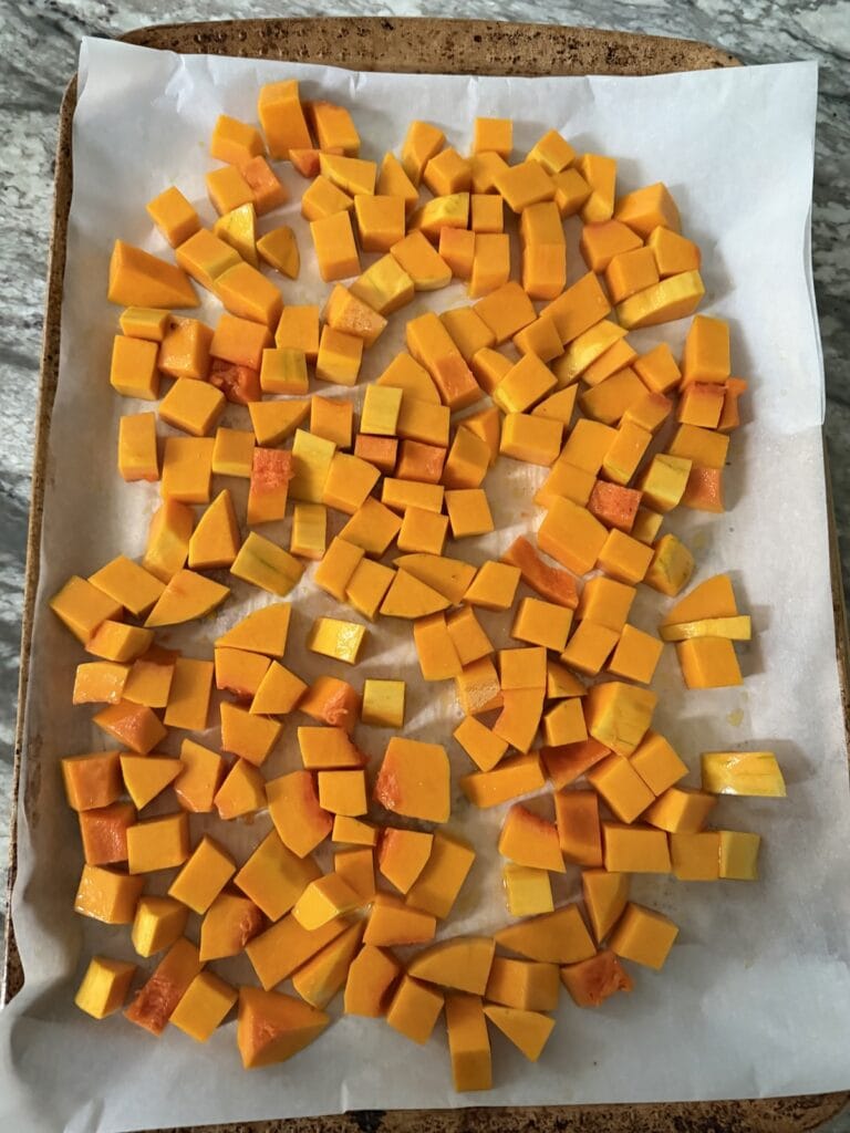 Cubed squash piecesd spread out on parchment-lined sheet pan, ready to roast.