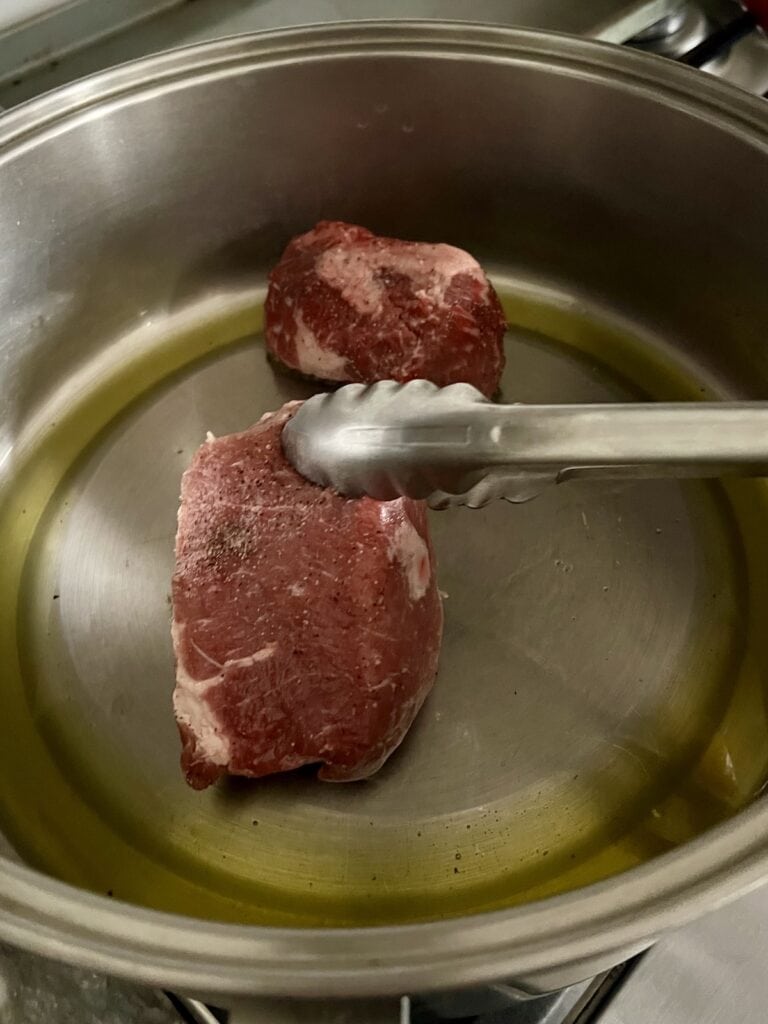 Using tongs to place a piece of meat into pan with oil to brown.