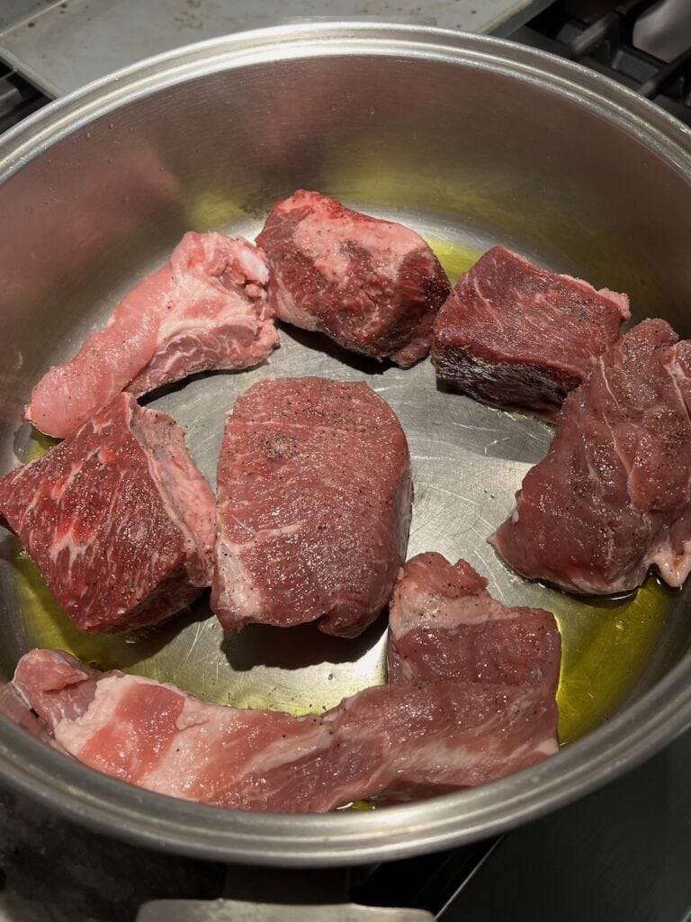 Multiple pieces of beef and pork browning in oil in large pot.