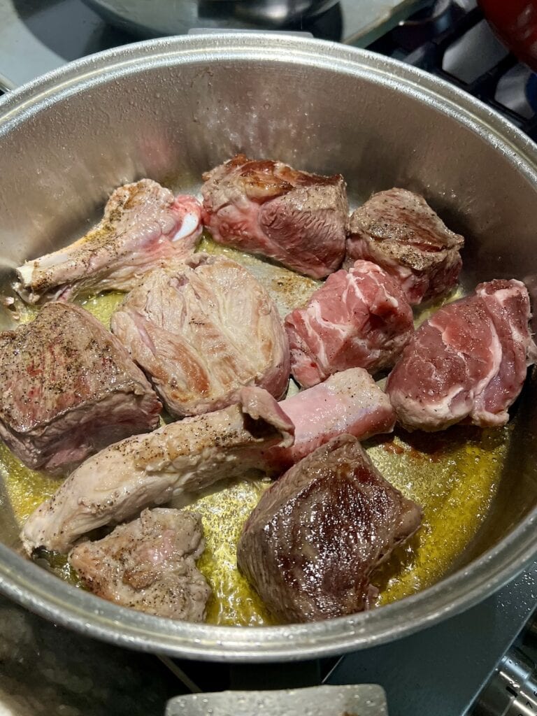 Multiple pieces of beef and pork browning in oil in large pot for italian american sunday gravy.