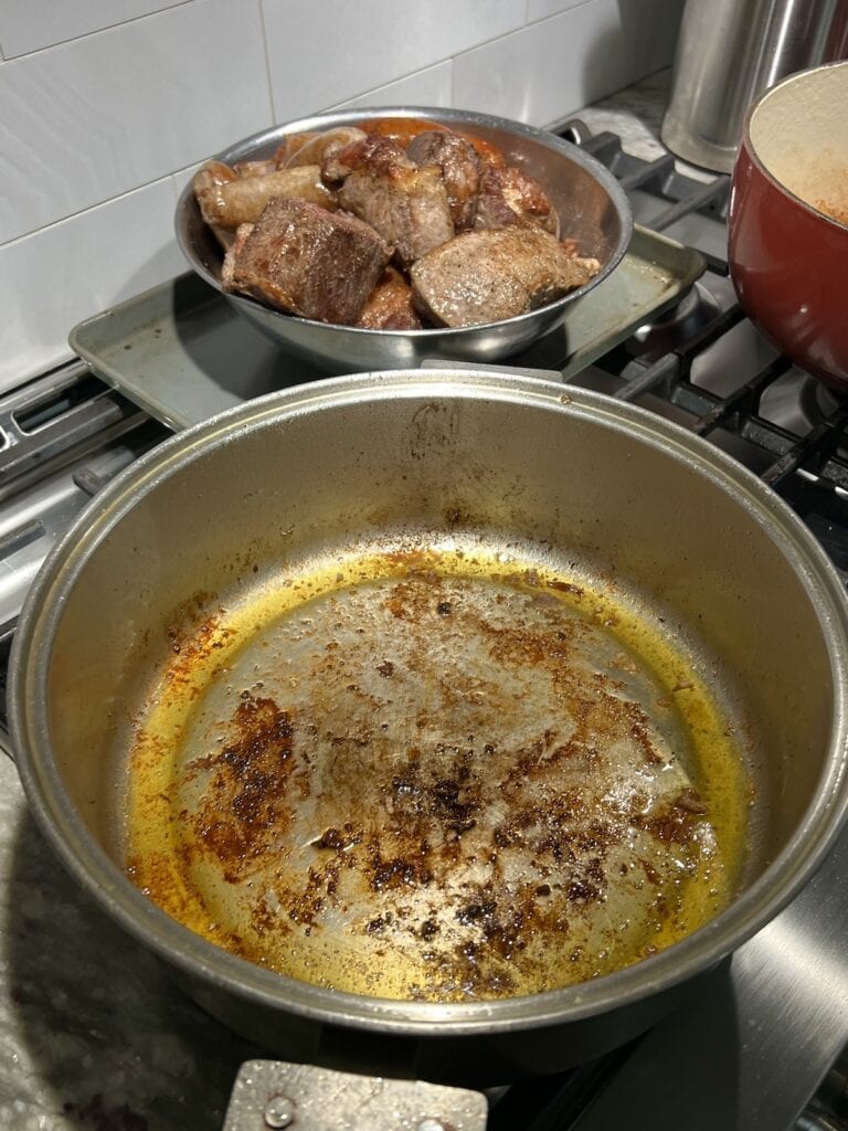 Empty pot filled with finds after searing all meat, with bowl of meat in the background.