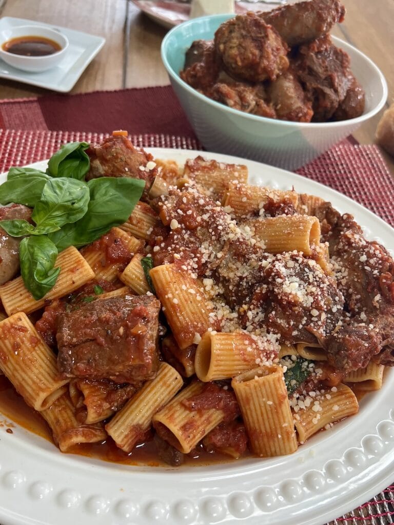 large platter of sunday gravy and basil garnish with grated cheese with bowls of meat and spicy oil in background.