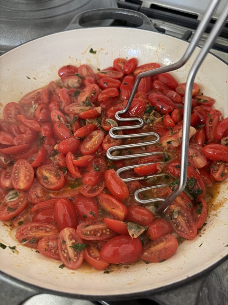 Lightly smashing tomatoes in pan with a potato masher.