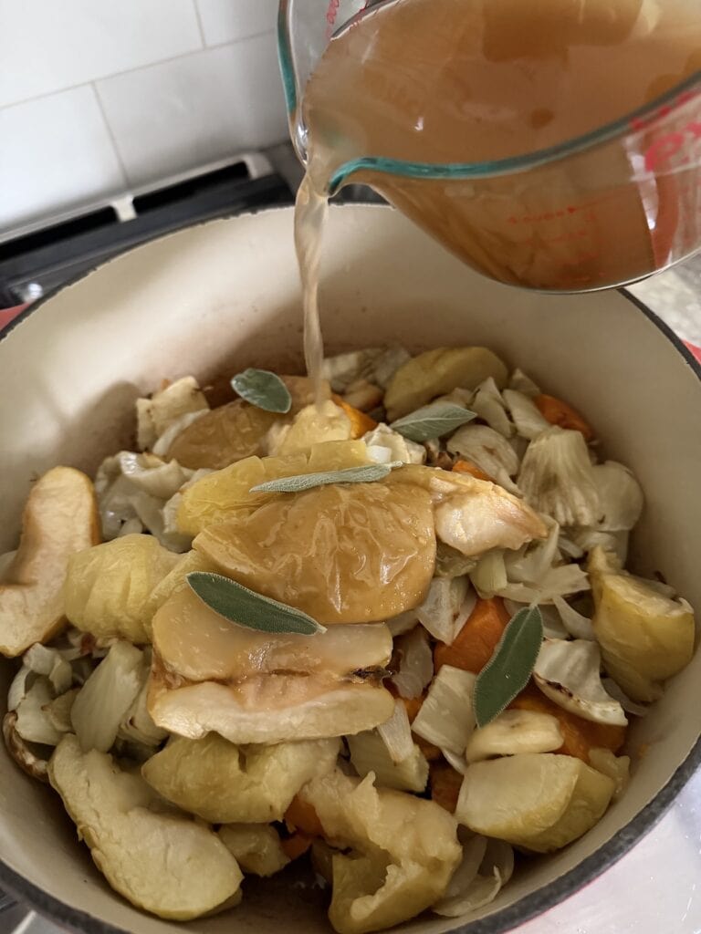 Pouring vegetable broth into pot of roasted vegetables and apples with sage leaves.
