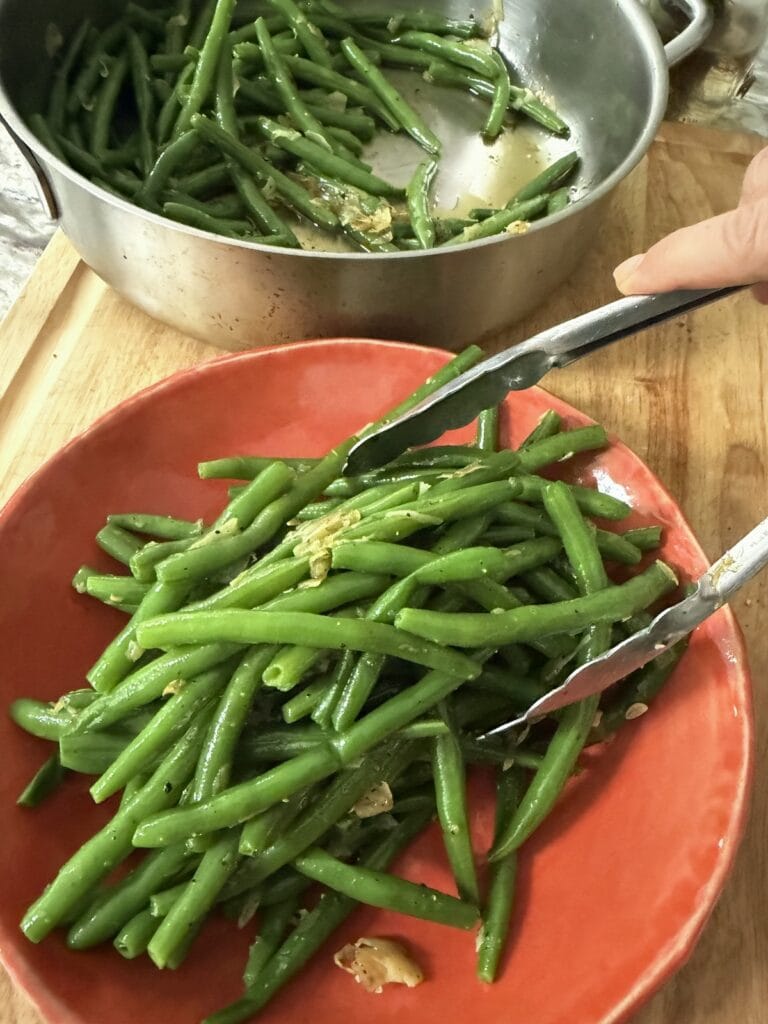 Using tongs to transfer Garlicky Sauteed Green Beans from sauté pan to orange serving bowl.