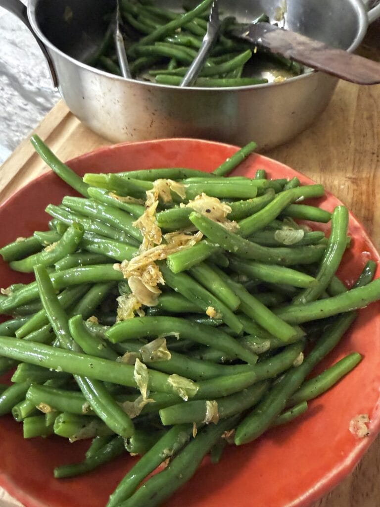 Garlicky Green Beans topped with sauteed garlic in orange serving bowl in front of pan of the veggies.