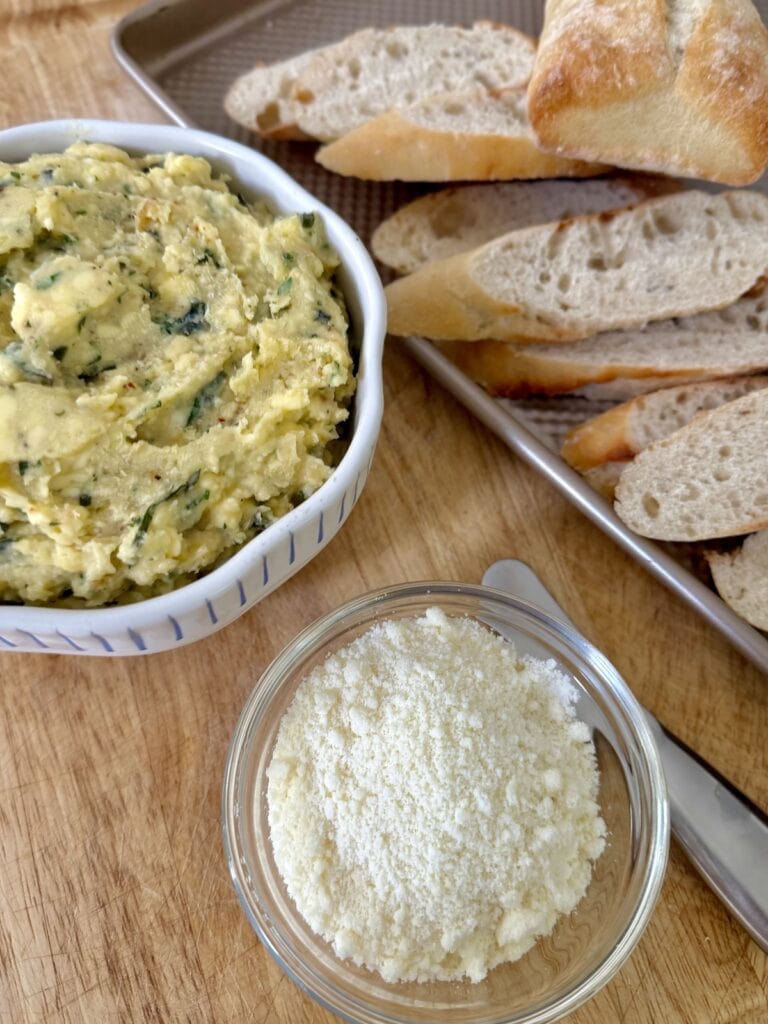 Bowl of garlic butter, bowl of grated pecorino cheese and slices of baguette on cutting board.