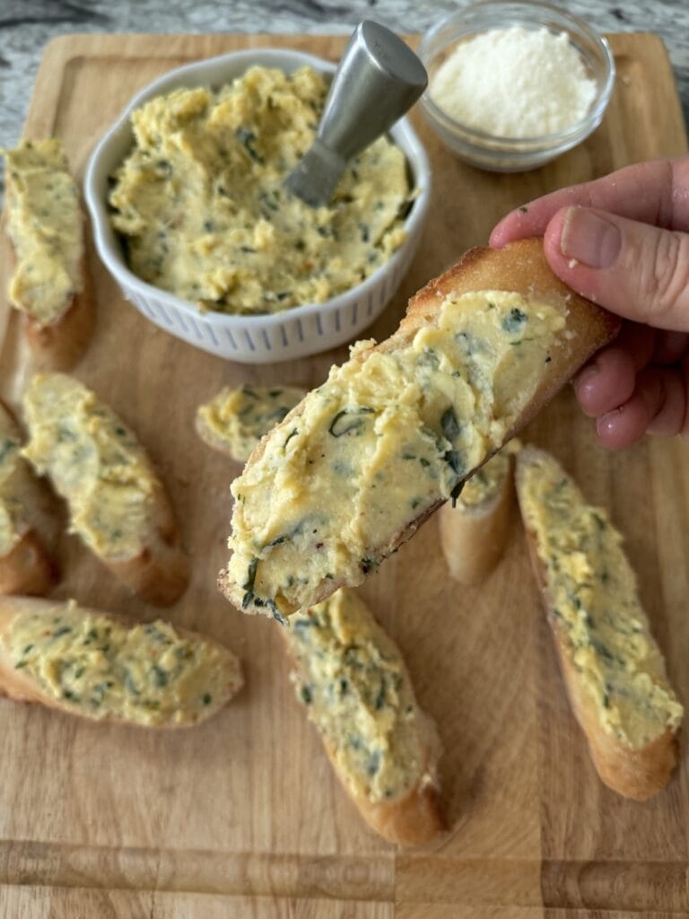 Buttered baguette slices with bowl of garlic butter in background.