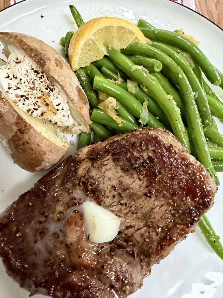 Italian Sautéed Green Beans with lemon wedge, seared steak and baked potato on dinner plate.
