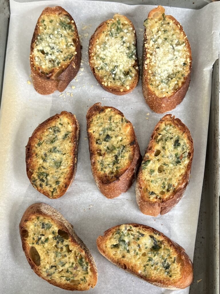 Several slices of homemade garlic bread baguette on sheet pan after broiling.