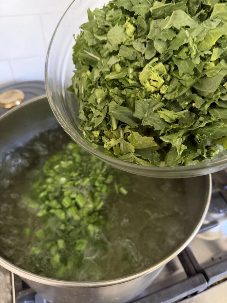 Adding chopped rapini leaves and flowers to boiling water in pot.