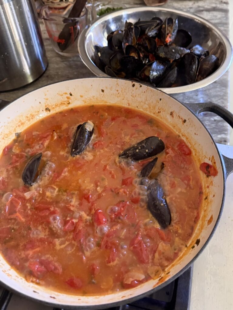 Four mussels left cooking in tomato sauce with bowl of cooked mussels next to it.
