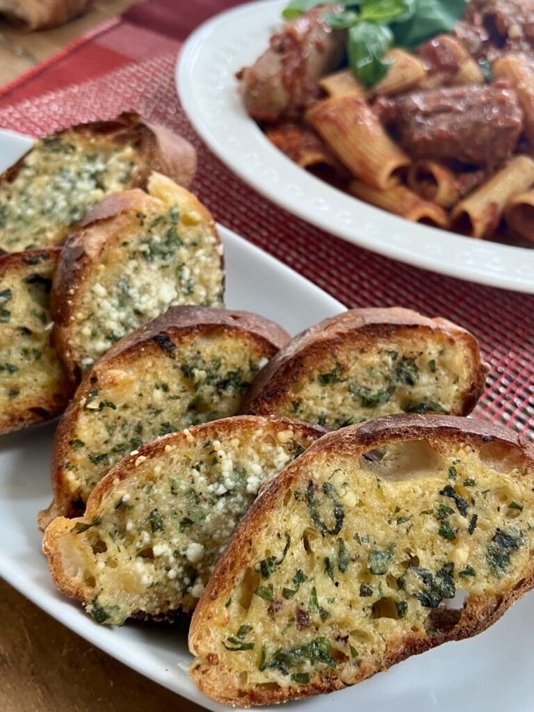 Slices of garlic bread on white platter with bowl of pasta in background.