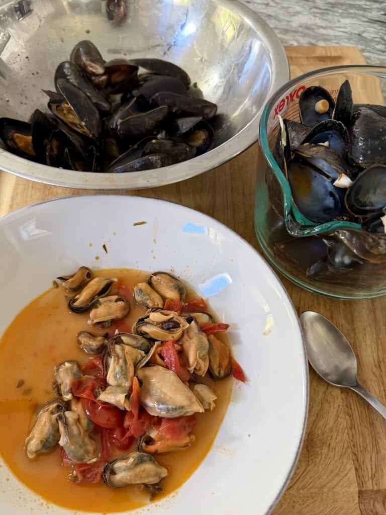 Shelled mussel meat in bowl next to shells and a bowl of some mussels still in shells in background.