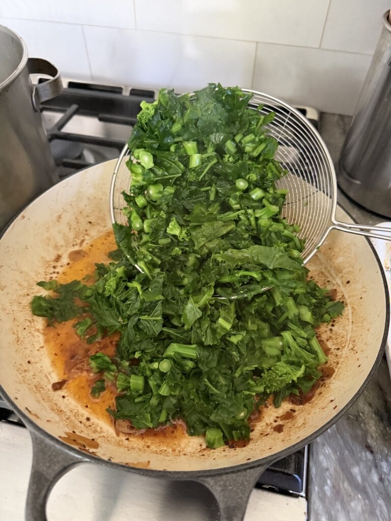Transferring cooked broccoli di rape to sauteed garlic and wine in pan with a spider scoop.
