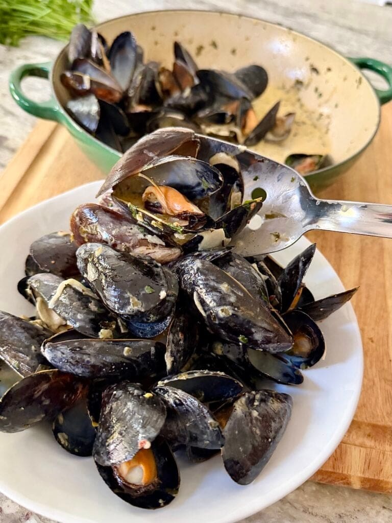 Spooning mussels with creamy white wine sauce from cast iron pan to serving bowl with stainless steel spoon.