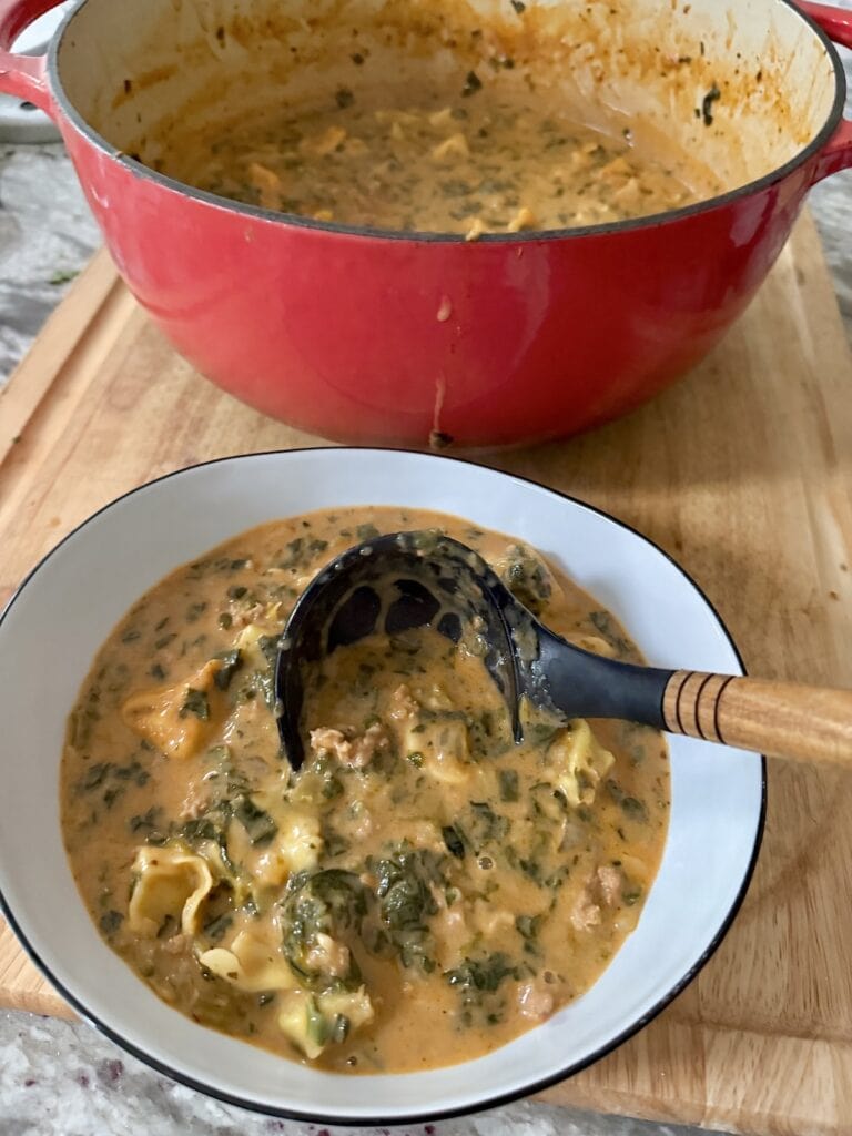 Ladling tortellini sausage soup into soup bowl from Dutch oven filled with it.