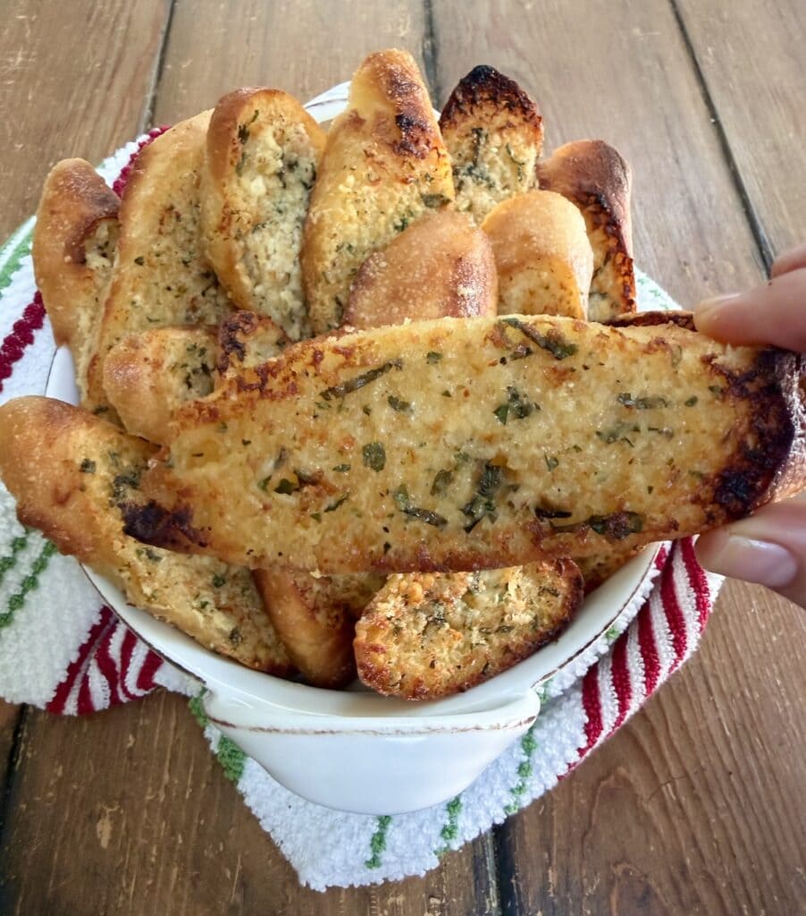 Piece of garlic bread baguette in hand over a bowlful of it.