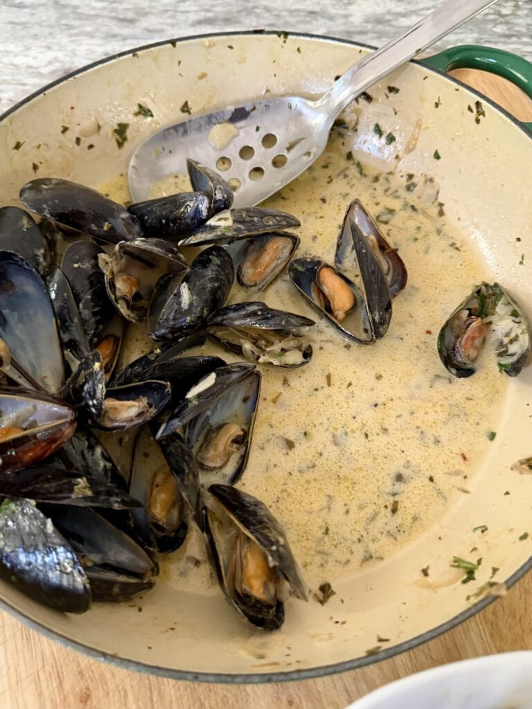 Cooked mussels in creamy white wine sauce with spoon in pan.