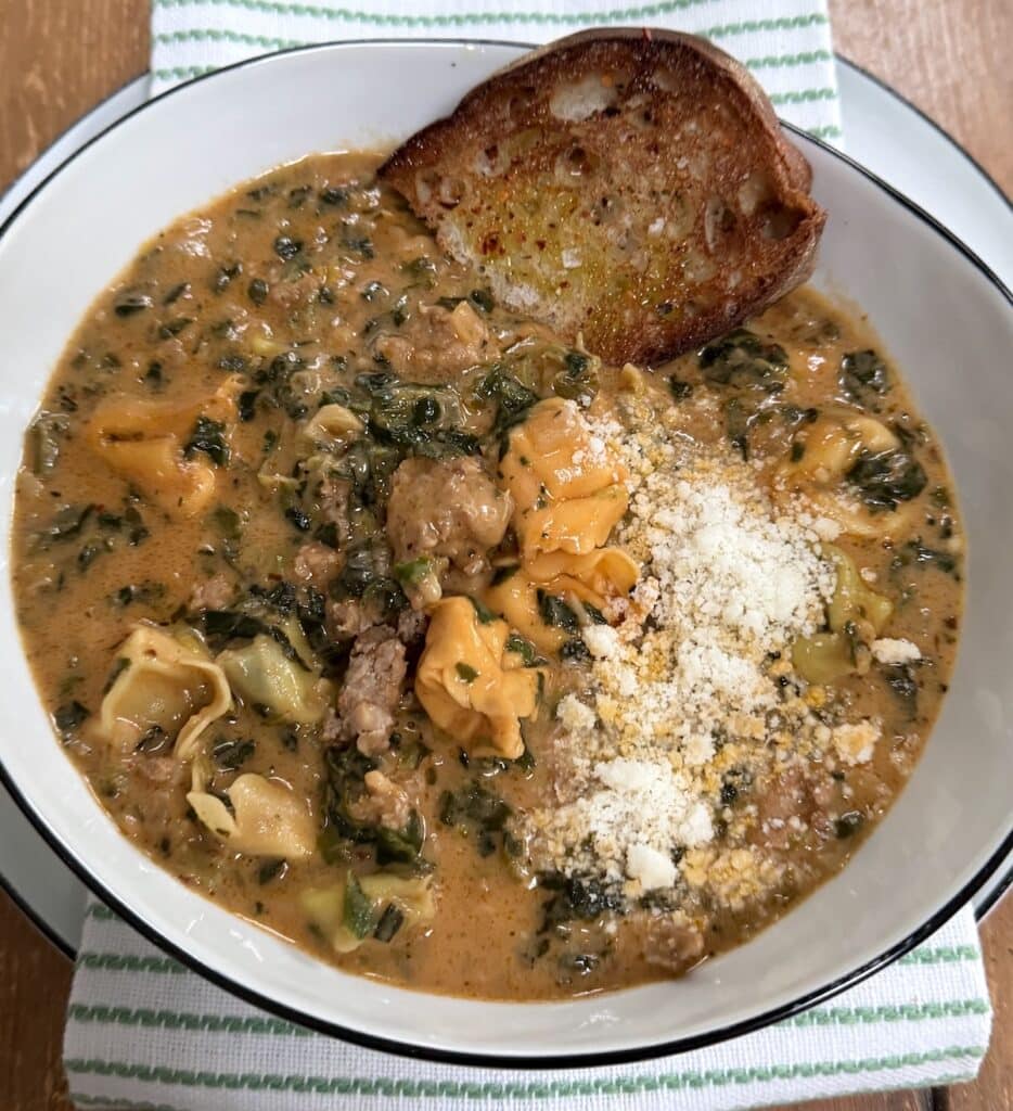 Sausage tortellini soup with grated cheese in large bowl with piece of bread over decorative towel.