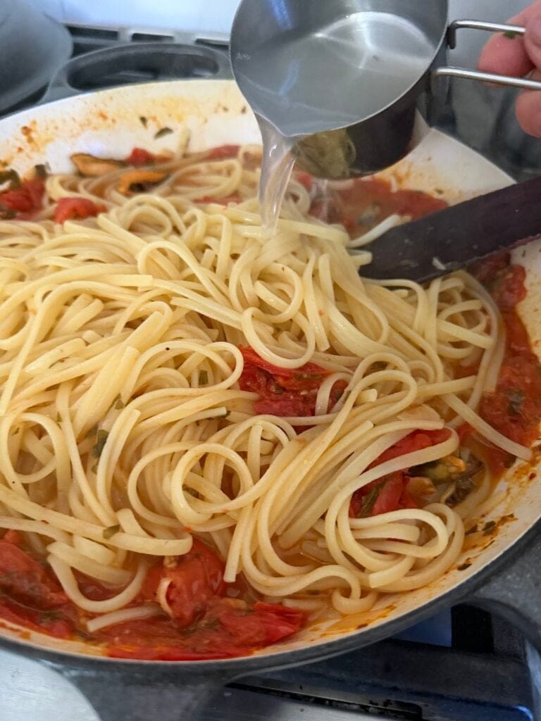 Pouring some pasta water into pan with cooked pasta and sauce.